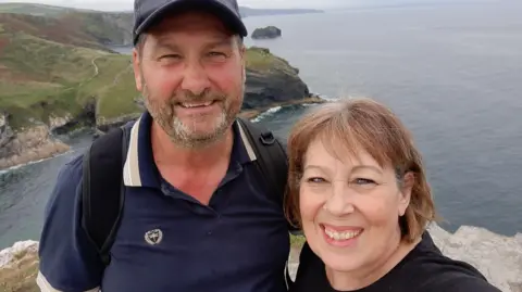 Lynda Sawyer  Kim and Lynda Sawyer smile for a selfie as they stand on cliffs with a view of the sea and coastline behind them. Kim, who has a beard, is wearing a dark blue polo shirt, a dark blue baseball cap and a black backpack. Kim, whose hair is cut in a bob, is wearing a black top.