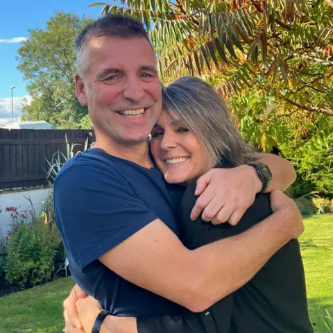 Jeannie Ambrose A couple - Jeannie Ambrose and her husband - are hugging in a garden. They both are smiling at the camera.