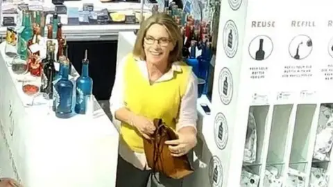 Norfolk Constabulary Gaynor Lord on CCTV dressed in a white shirt, with a yellow tank top and is carrying a brown bag. She has blonde hair and glasses and is pictured at work at a gin counter.