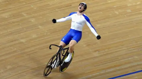 PA Chris Hoy in white and blue kit with his arms outstretched victoriously as he rides a bike