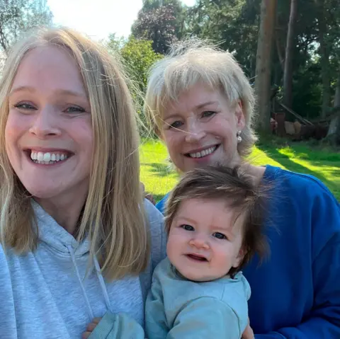 Sally Magnusson  Selfie of Sally Magnusson (right) in a navy blue top next to her daughter Anna Lisa who is wearing a grey hoodie. Anna Lisa is holding her daughter Remy who has dark hair. The sun is shining on the bright green grass and trees behind them and they are all smiling at the camera.