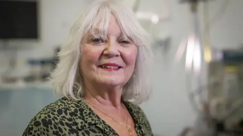 BBC Doreen Adams, woman with shoulder-length grey hair and middle parting, smiling, wearing a green and black blouse and a necklace, with medical equipment in the background.