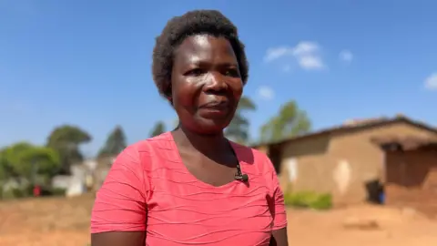 BBC Dorothy Masasa standing outside in a pink t-shirt in the sun. There is a single storey building in the background.