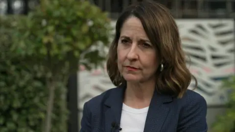 BBC A medium close up of Pensions Secretary Liz Kendall wearing a blue jacket and white top with tree and bushes in background