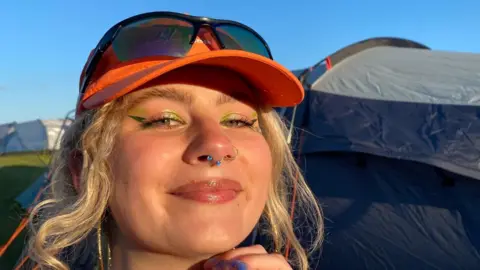 Imogen Turnbow A woman wearing an orange hat and with colourful eye make-up smiles with the sun shining on her face