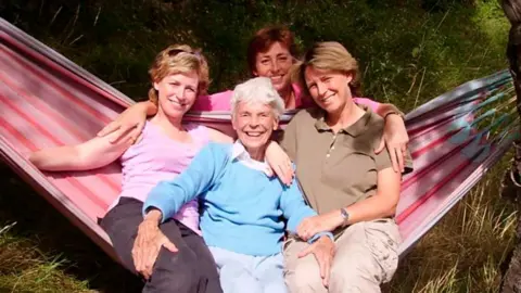 Sally Magnusson  Sally Magnusson (left) in brown trousers and a pink T-shirt with sunglasses on her head, with her mother Mamie (centre), wearing a light blue jumper with white blouse underneath, sister Margaret (right) in beige trousers and a brown T-shirt. The three woman are sitting on a red and pink striped hammock with grass around them. Sally's sister Anna is crouching down behind them and is wearing a pink long-sleeved top.