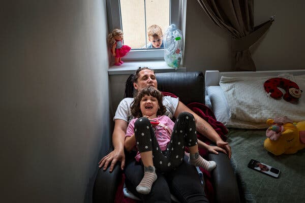 Sonya, a 5-year-old with a mop of dark hair, on top of her mother in an armchair, her lighter-haired brother watching from outside.