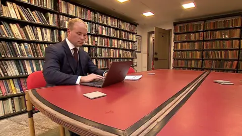 Tom Longstaff sits in a smart suit at a desk concentrating at his laptop 