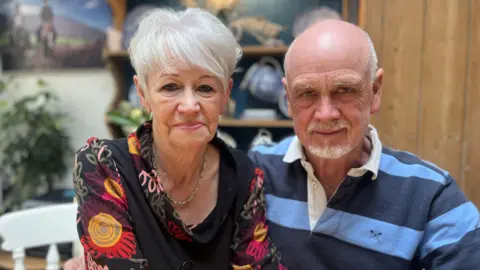 A man and a woman sit looking into the camera. The woman has short grey hair and is wearing a gold necklace and a black top with purple, yellow and red flowers on it. The man is balding with short white hair at the sides and a white goatee beard. He is wearing a blue striped polo shirt. Behind them is a plant, and a wooden cabinet with crockery on display.