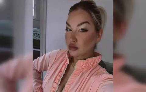 Beautician Sarah Guy in a selfie photograph wearing a coral pink frilly blouse with a plunging neckline. She has large painted lips and arched eyebrows.