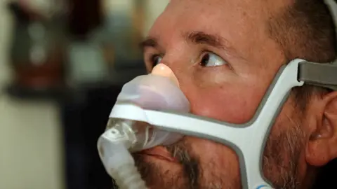 BBC A close up of Nik Ward, an MND sufferer, sitting in his motorised wheelchair while he looks towards a family member standing to the left of the camera. He has a breathing apparatus across his nose.