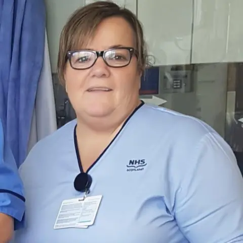 Susan McGowan looks into the camera smiling. This is an older photo of her, taken at Monklands hospital while she is wearing her nursing scrubs and ID badge. She wears her hair back.