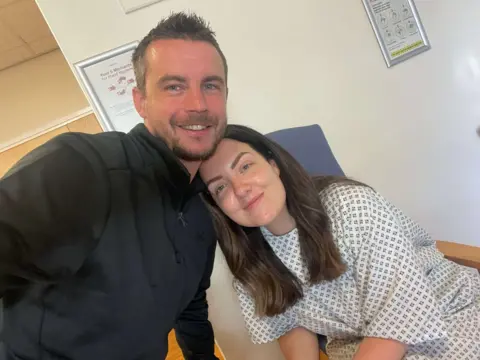 Anna Cooper Photo of lady in hospital gown in a clinical area of a hopsital with her partner, a man dressed all in black. Both are smiling at the camera. 