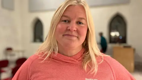 BBC Jo Broughton wearing orange/pink towel top inside a large room