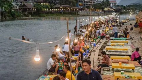 Getty Images People drink and enjoy food on a riverbank in the town of Vang Vieng in Laos. There as some people swiming in the river and a string of lights highlights dozens of people sat on tables.