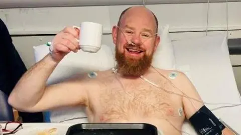 Richard Donaldson Man with ginger beard in hospital bed, with bare chest and hooked up to wires, smiling, and holding up a white mug by its handle in a 'cheers' gesture.