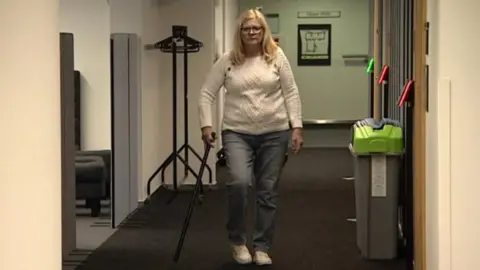 A woman in a white jumper, blue jeans and white trainers holding a walking stick while slowly walking down a corridor with a slight limp.  The corridor has dark grey carpet and white walls with a black metal clothes hanger on one side and what look like grey bins with green and blue lids on the other side.