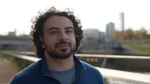 A head and shoulders portrait of a doctor with dark curly hair and facial hair, wearing a blue top. He is photographed outdoors with an urban skyline visible in the background. He has a thoughtful expression and is looking directly at the camera