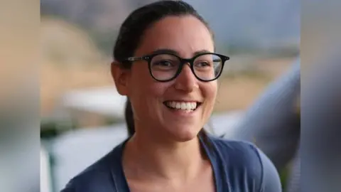 Martino Brizio A young woman with brown hair, brown eyes, wearing dark framed glasses and wearing a mid blue top smiles looking slightly off camera, the background is blurred.