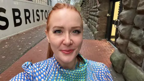 Anna Hill Anna Hill smiling while stood outside. She has ginger hair in a ponytail and is wearing a bright blue dress with dots.