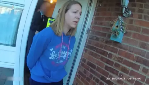 Cheshire Constabulary Lucy Letby shown on police body cam being arrested and taken from her home. She is wearing a blue Lee Cooper hoody and is walking past a brick wall with a bird box on it. The date read 2018/07/03 and the time is 06:14:40. She is followed by a police officer and a plain clothes detective who are blurred out. 