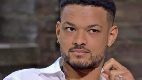 BBC Steven Bartlett pictured on the set of Dragons' Den. He has short afro hair with a fade on the side, and a short beard. He is wearing a white, open necked shirt, small hooped and stud earrings, a gold chain necklace and a gold ring studded with coloured gems. Behind him is an exposed brick wall. 