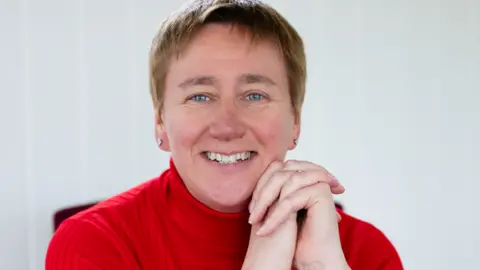 Jenny Smith Photography Diane Danzebrink is smiling at the camera while wearing a red turtleneck top and silver stud earrings. She has light brown short hair and blue eyes. She has her elbows on the table and hands clasped upward in front of her, so her chin rests on them.