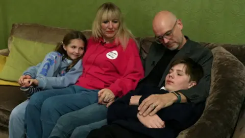 A family of four sit on a green sofa. The mother and father are sitting in the middle with their daughter sat on the left and their son sat on the right. 