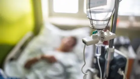 Getty Images A stock image showing an out-of-focus patient lying in a hospital bed. In the foreground and in focus is a drip.