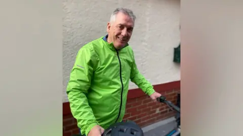 Kevin Hill wearing a fluourescent green jacket and holding his bike and a helmet and smiling.