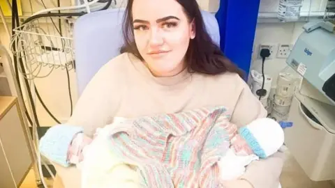 Meg Hughes Meg Hughes, a woman with dark hair, smiles into the camera while holding new-born twins in her arms. The babies are wrapped in a rainbow-coloured blanket and each wearing woollen hats with tubes in their noses. The picture is taken on a hospital ward.