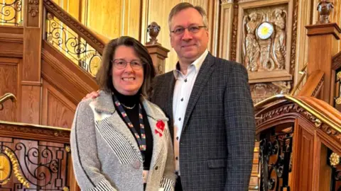 Peter has short grey hair and round glasses and is wearing a dark blazer and white shirt. Jill has dark shoulder length hair and large round glasses. She is wearing a grey woollen blazer with black and white stripes and a black top underneath. They are standing in front of a wooden staircase and are looking at the camera, smiling.