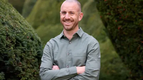 Paul Galdas A man in a grey shirt, beaming, stands with his arms folded in front of bushes and an area of woodland