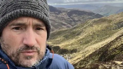 Dan Somers A selfie of a man with a woolly hat on in front of undulating hills