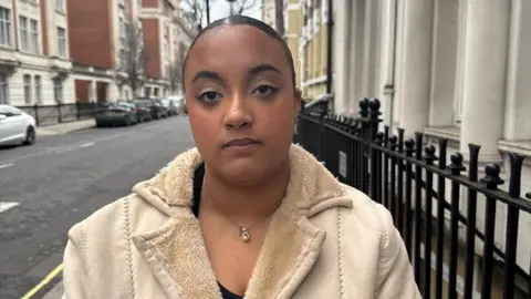 A young woman with pulled-back black hair. She is wearing a white jacket with woollen lapels. She is standing on the pavement next to some black railings