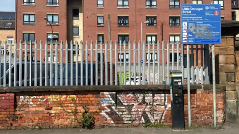 The sign for Little Donegal Street car park. The wall behind is red and is covered in graffiti. There are tall spiked metal gates surrounding the car park.