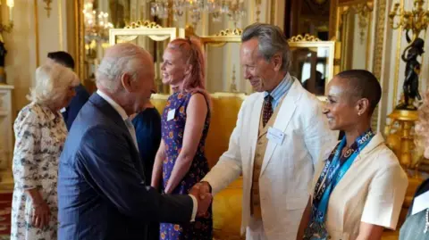 PA Media King Charles shakes hands with Richard E Grant at a reception at Buckingham Palace. Adele Roberts stands to Grant's left. Queen Camilla is greeting guests behind the King.