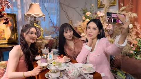 Three young women with surgically-enhanced faces, having tea and cakes together, pose for a selfie 