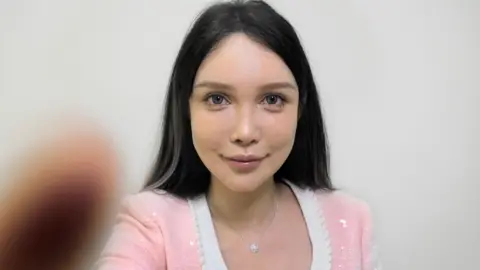 BBC A young woman with a surgically-enhanced face, wearing a pink jumper, stares at the camera. White lines have been imposed over her face by a cosmetic surgery app called SoYoung 