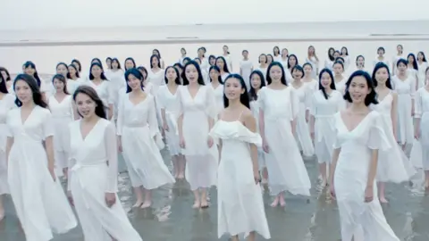 SoYoung A huge crowd of attractive women in white dresses are stood bare-foot on the soft sands of a beach, staring at the camera
