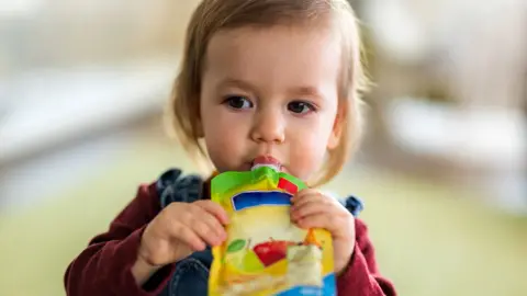 Getty Images A toddler with blondey-brown hair sucking a pouch of baby food, which she is holding with both hands. She is wearing a burgundy top and dungarees. 