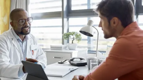 Getty Images A man is sat at a doctor's clinic, looking at the doctor, who is speaking to him at a desk and has a notebook open
