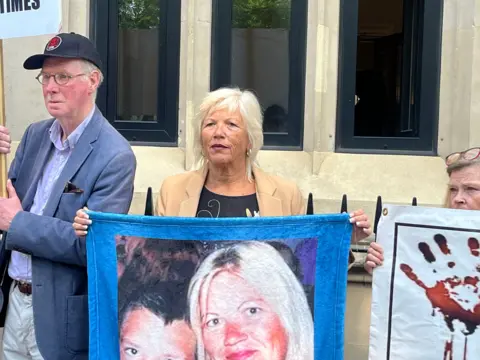 RICHARD DANIEL/BBC Melanie Leahy outside the inquiry. She is holding a picture of her with her son, Matthew. She has shoulder-length blonde hair and is wearing a sand-coloured jacket. Other protesters are either side of her.