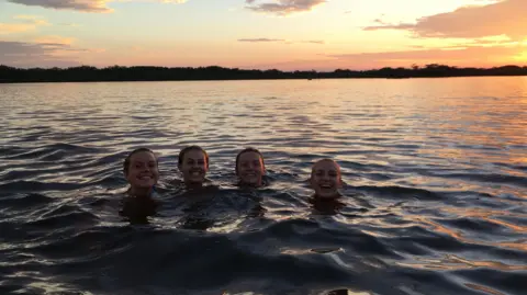 Ellie Irwin Ellie and friends swimming in the Amazon river, with their heads above the surface and the sun rising behind the trees in the background.