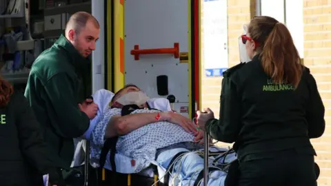 EPA Two London ambulance staff, wearing green clothes, wheel a patient on a trolley, with an ambulance in the background