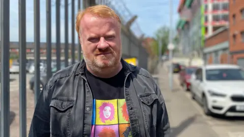 BBC Brian Smyth has short, combed over red hair and a red and grey beard. He is standing next to the gates of a car park and is wearing a dark denim jacket and a black t shirt with a multicoloured pattern.