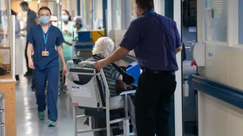 PA Media A view in a hospital corridor, where, in the foreground, one member of staff stands in front of a chair where an elderly patient sits. In the background, a female member of staff walks down the corridor towards the camera.