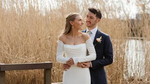 Ellie Irwin Ellie and her husband stand together in their wedding outfits on their wedding day, holding hands and staring into each other's eyes