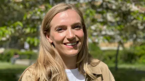 BBC Dr Ellie Irwin faces the camera, smiling, with long blond hair in a sunny outdoor place with blurred trees in the background