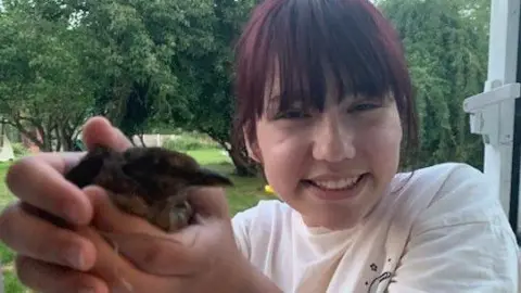 Family handout Elise Sebastian holds a small bird in her hands. She is smiling happily and is wearing a white T-shirt. Elise has dark brown hair with a fringe which is dyed red. She is outside with trees in .